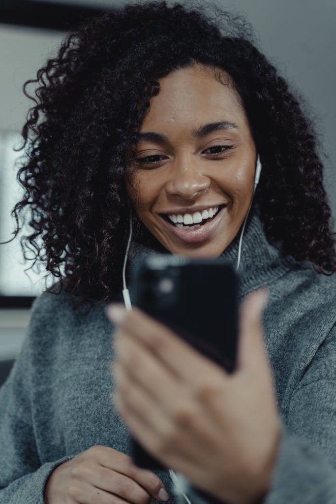 A joyful woman in a gray sweater uses a smartphone with earphones indoors.